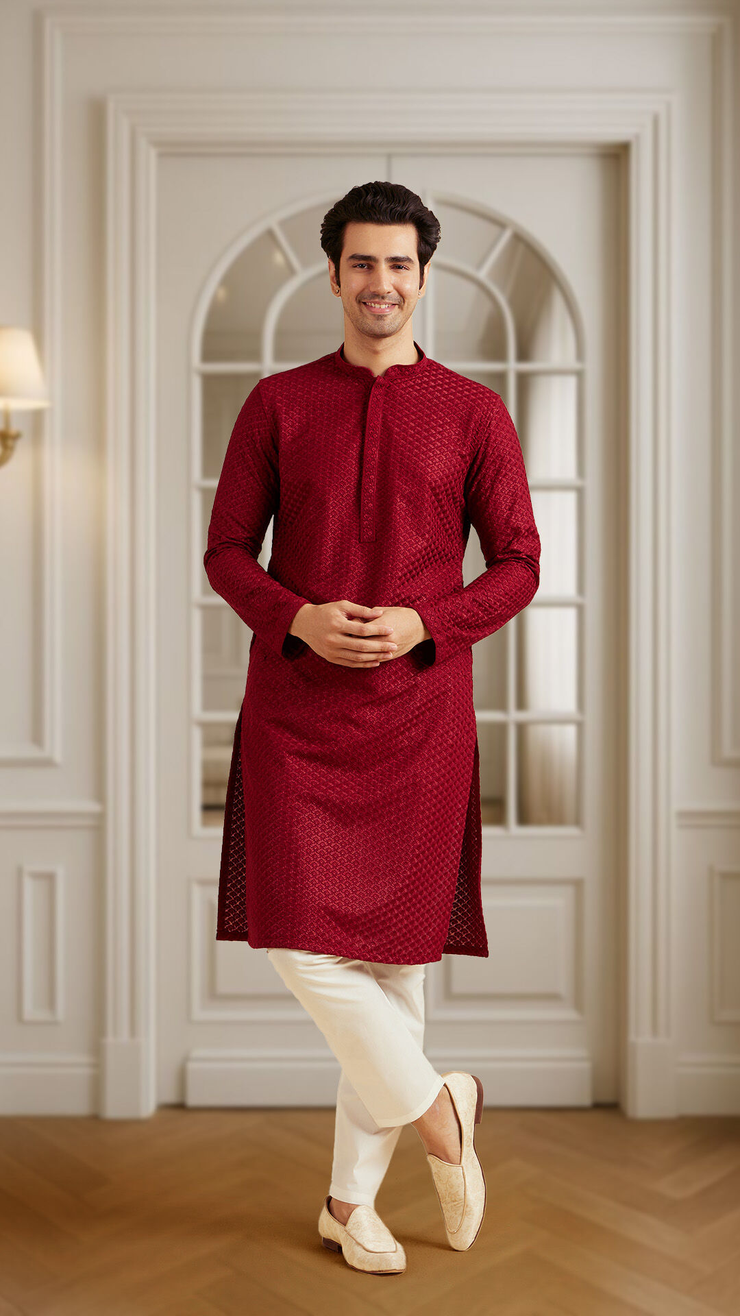 Man dressed in a maroon Chikankari embroidered kurta paired with white churidar pants and traditional mojaris, posing with crossed legs in an elegant room with wooden flooring and arched window backdrop.